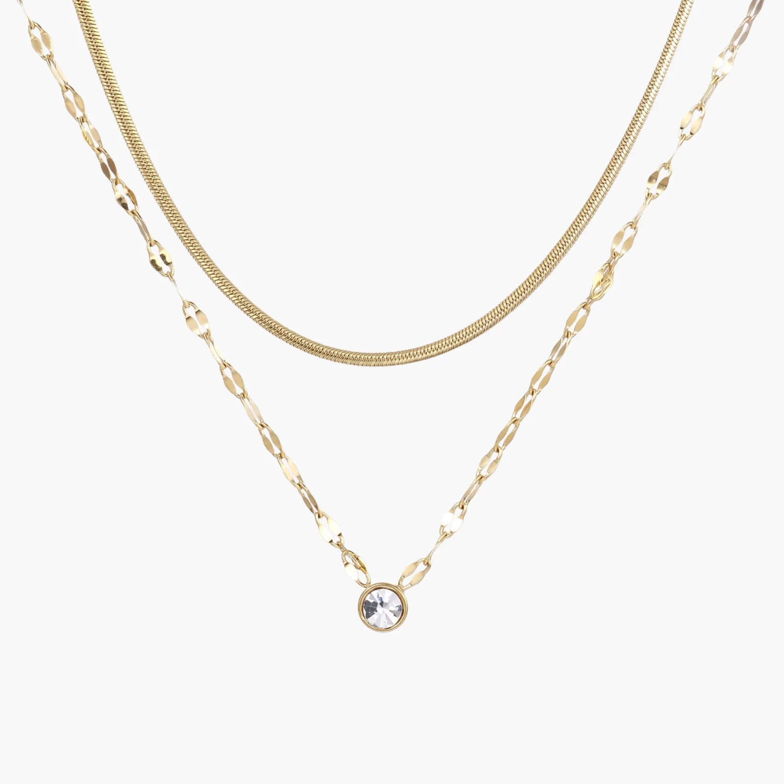 Layered gold necklace with herringbone and link chains, small bezel-set crystal pendant on cream background