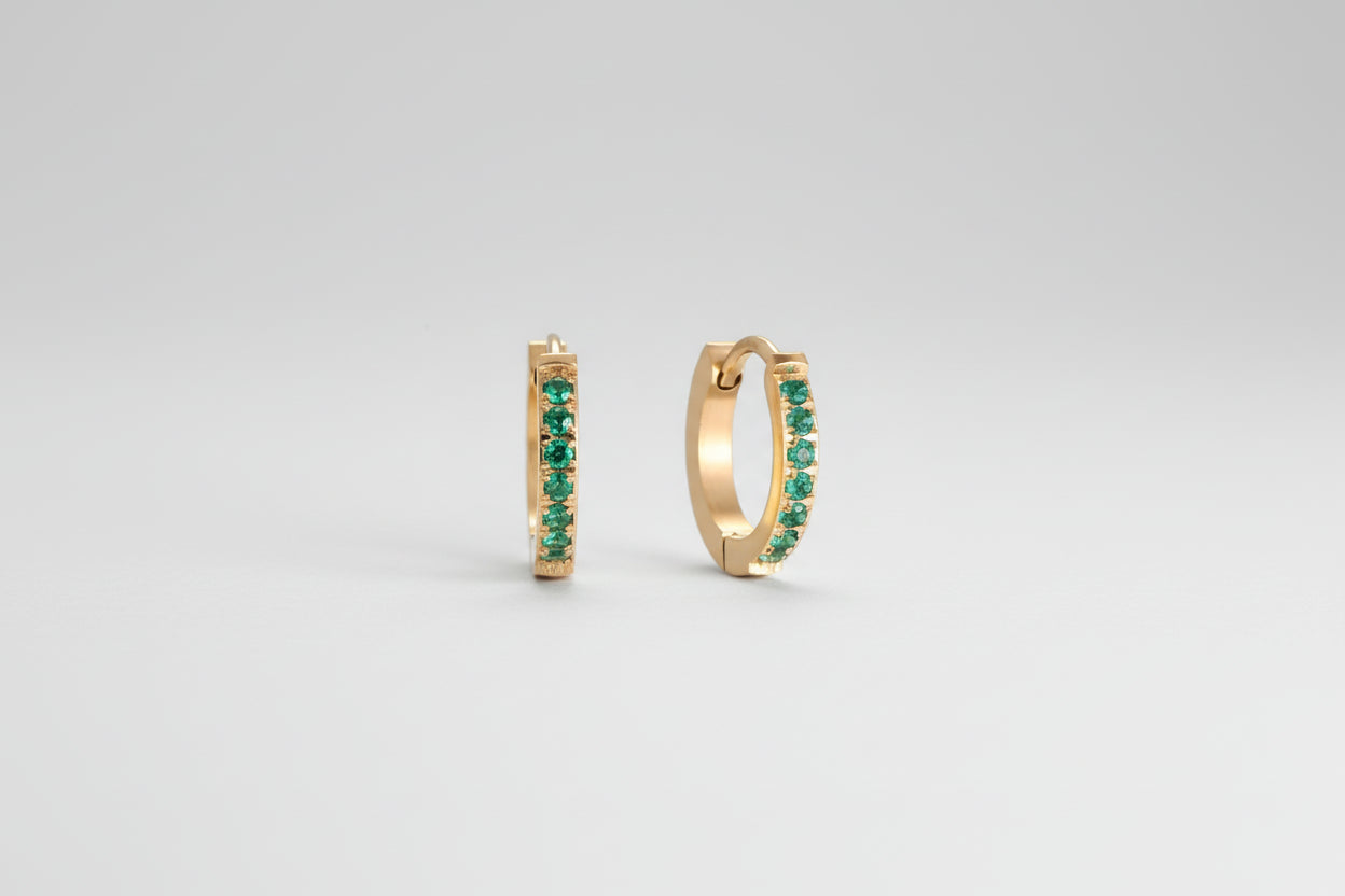 Pair of small gold huggie hoop earrings with bright green cubic zirconia stones on cream background